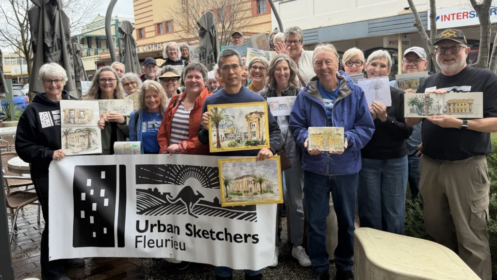 Members of Urban Sketchers Fleurieu smile behind their banner on a Victor Harbor street, holding up the sketches and watercolours they made that morning.