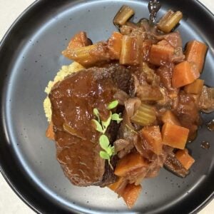 A glossy red wine braised beef short rib resting on wattleseed and pepperberry mash with braised carrots, celery and shallots, garnished with fresh thyme on a dark speckled plate