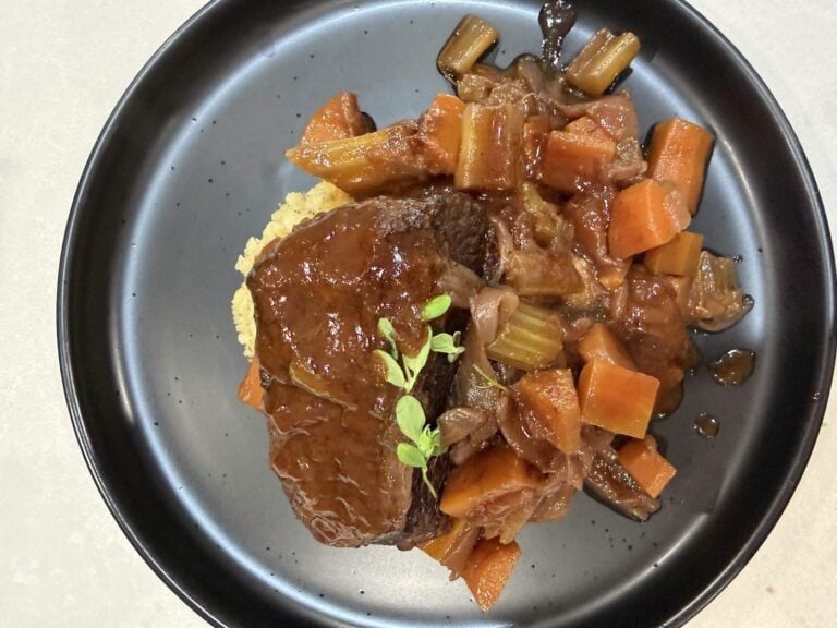 A glossy red wine braised beef short rib resting on wattleseed and pepperberry mash with braised carrots, celery and shallots, garnished with fresh thyme on a dark speckled plate