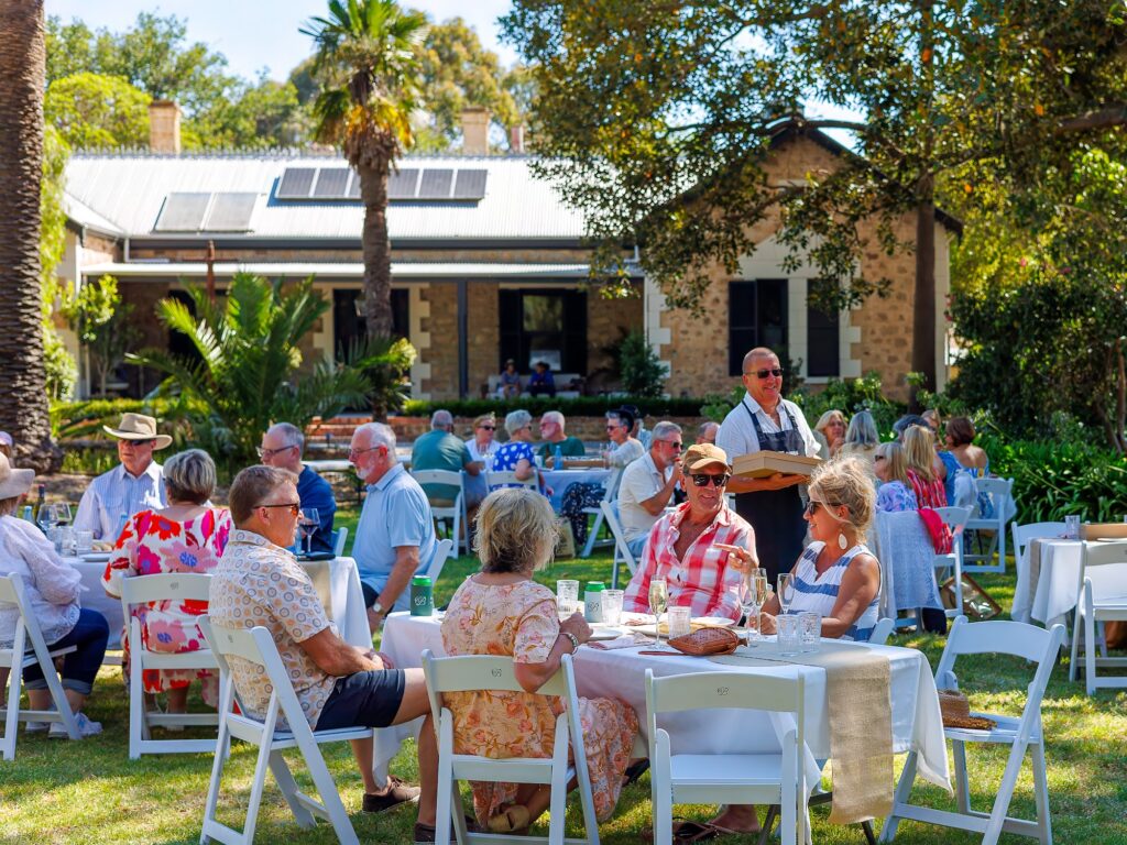 Guests sit at white-clothed tables on a sunny lawn under trees while a server carries a tray past a stone farmhouse.