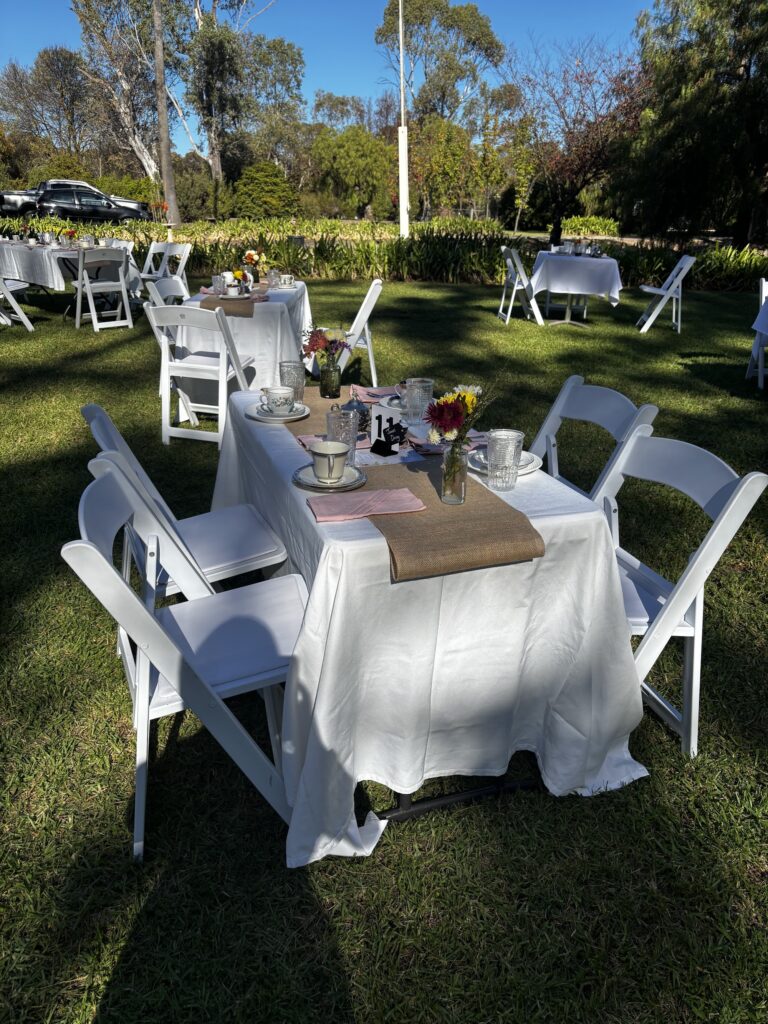 A white-clothed long table set with teacups, saucers, glassware and wildflowers on a burlap runner, on a sunny lawn.