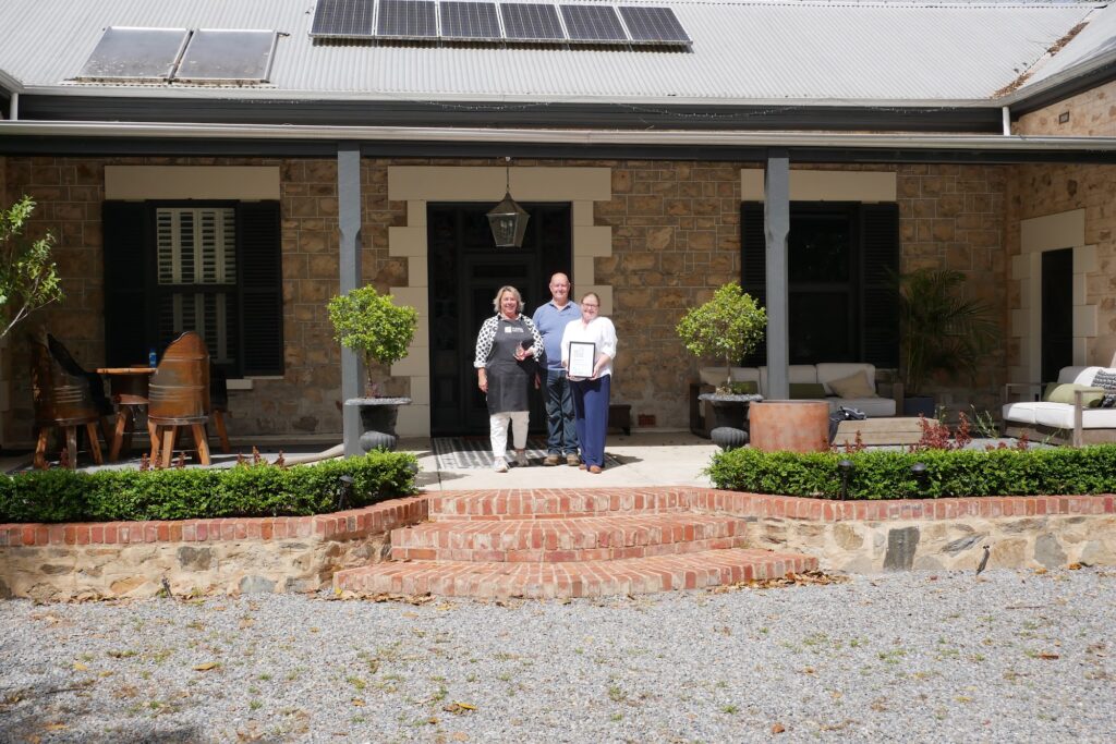 Three people stand smiling on stone steps outside a stone homestead, one holding a framed certificate.