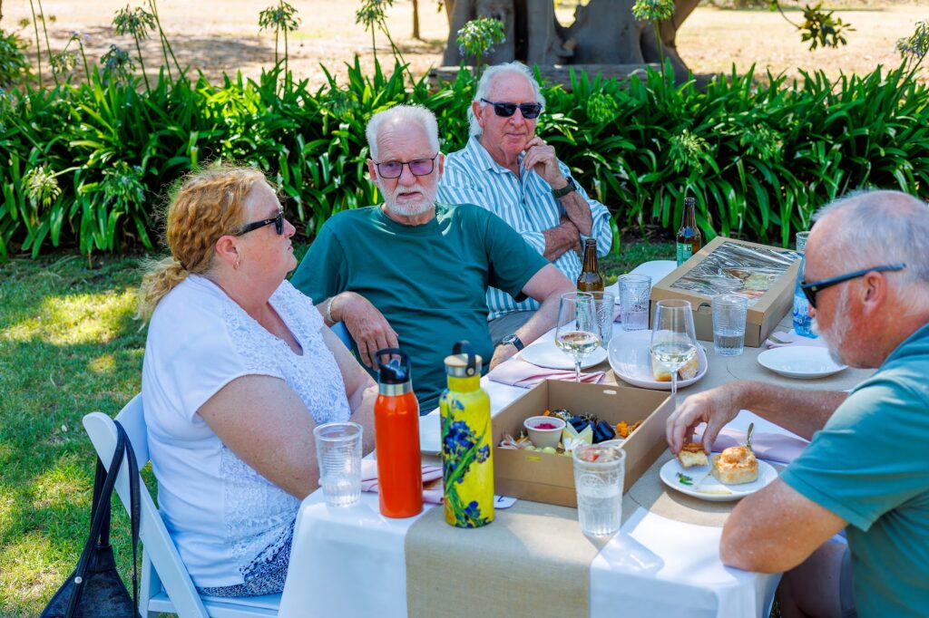Three older guests and one more man sit at an outdoor picnic table with wine glasses, bottles and platters of small bites.