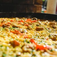 A close-up of chicken paella with red peppers and saffron rice, cooking in a wide pan at an outdoor market