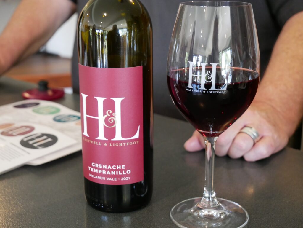 A bottle of red wine beside a filled wine glass on a grey bench, with a hand resting next to them.