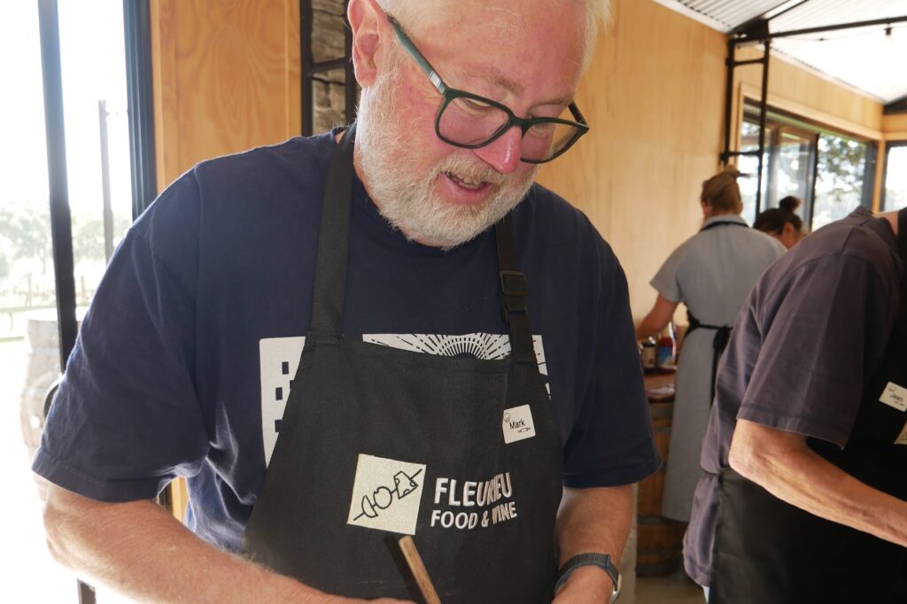 A man in a black apron stirs flour and egg in a metal bowl with a wooden stick while others work behind him.