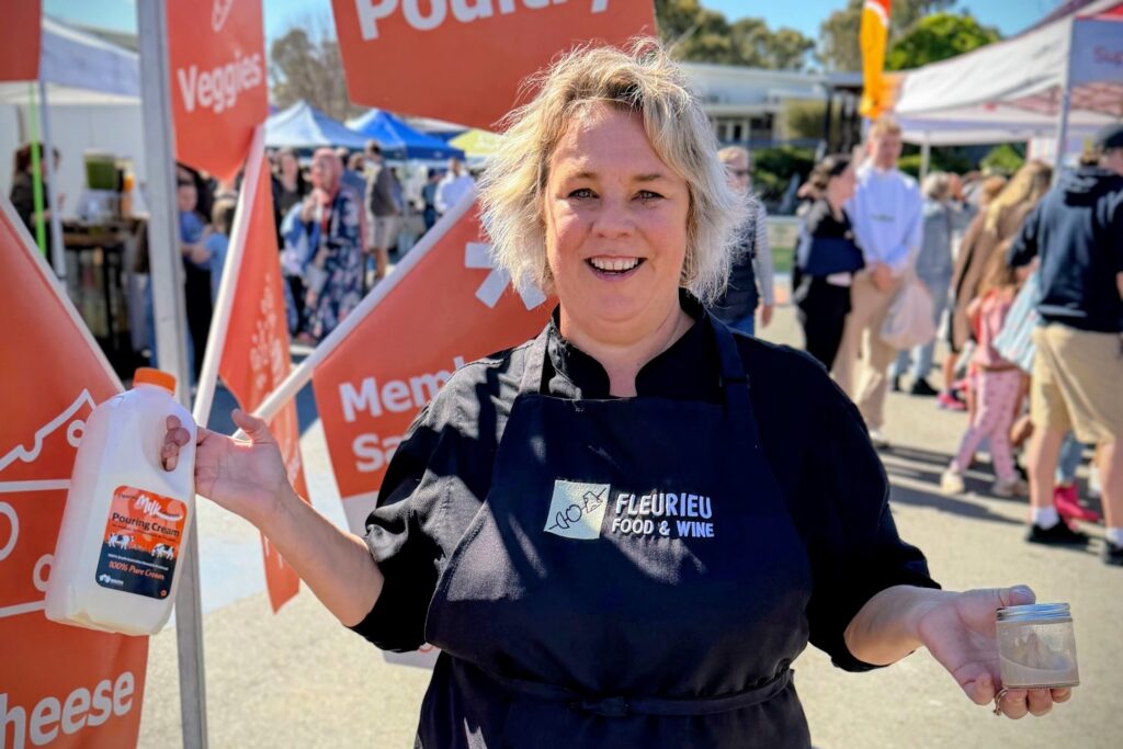 A smiling person in a black branded apron holds a bottle of cream and a small jar at an outdoor market with flags.