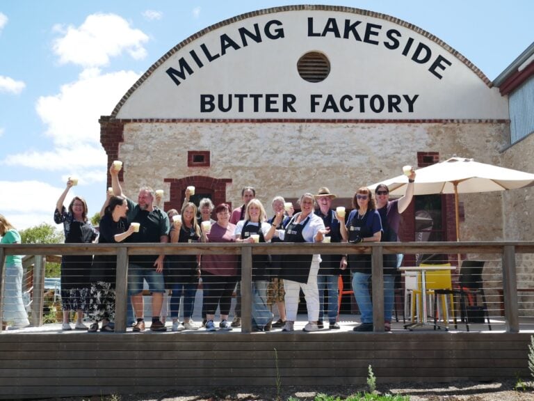 A group of smiling guests raise cheese curds and glasses on a timber deck in front of the Milang Lakeside Butter Factory.