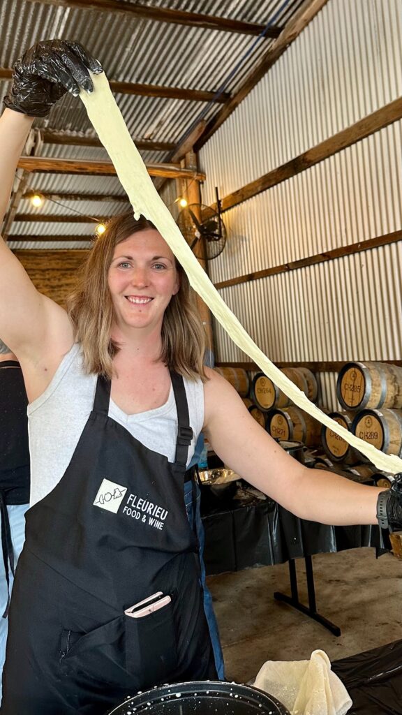 A smiling woman in a black apron and dark gloves holds up a long stretched ribbon of pale mozzarella curd in a rustic.