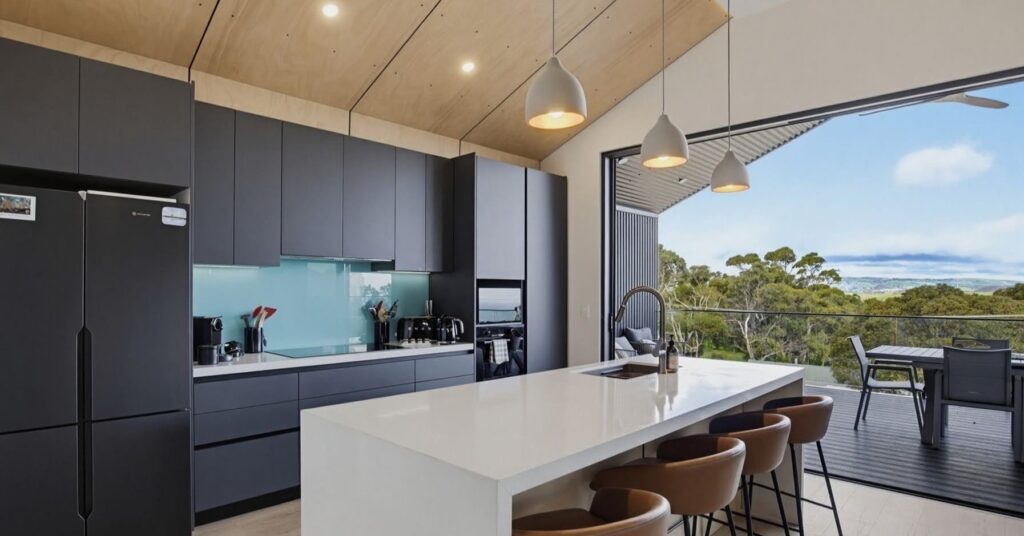 Modern kitchen with dark cabinets, a white island with bronze stools, and a wide view through glass doors to trees.