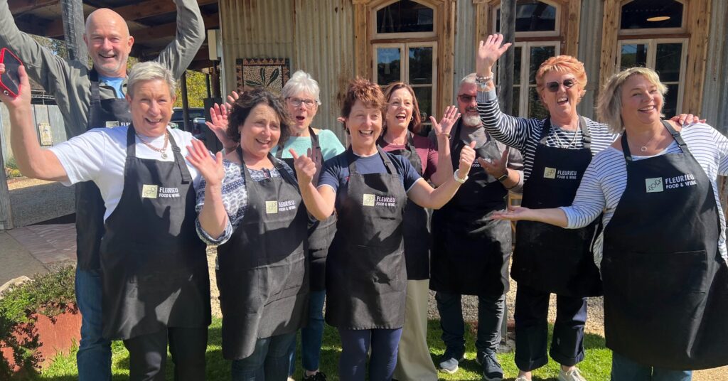 A group of guests in black aprons raise their arms and laugh on the grass after a Fleurieu Food and Wine cooking class on the Fleurieu Peninsula