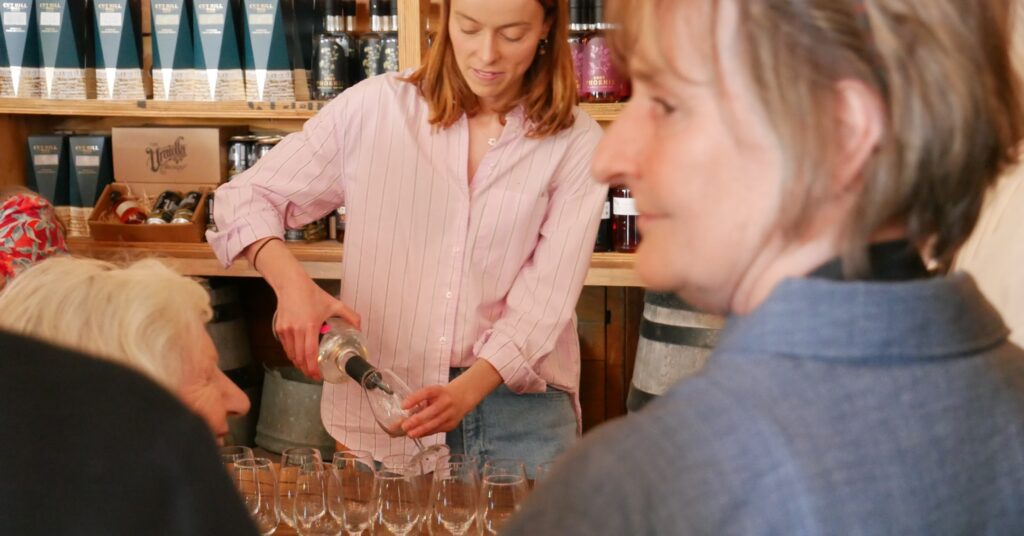 A woman in a pink striped shirt pours a clear drink into rows of tasting glasses as guests look on in a shop.