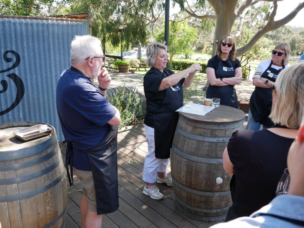 A presenter speaks to a small group of aproned guests gathered around wine barrels on a sunny outdoor deck.