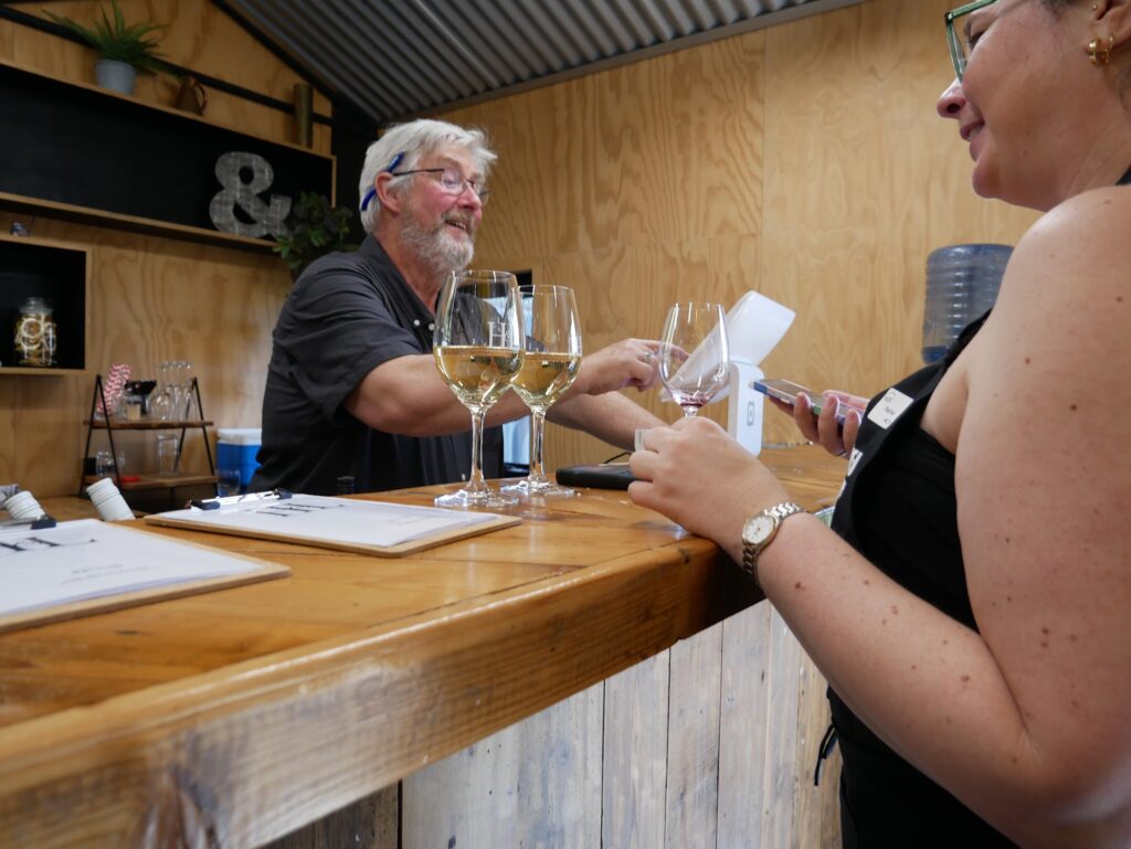 A man behind a timber tasting bar pours wine into glasses while a guest stands opposite holding a phone.