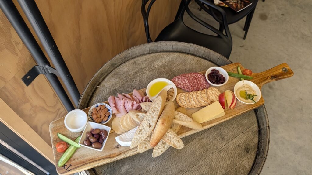 A long wooden board on a barrel table holding cured meats, cheese, crackers, bread, olives, nuts and dips.