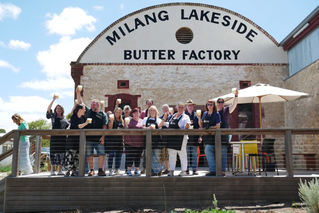 A group of smiling guests raise cheese curds and glasses on a timber deck in front of the Milang Lakeside Butter Factory.