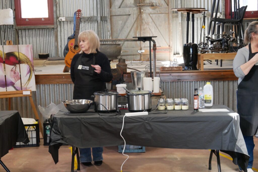 Presenter in a black apron speaking behind a draped table of pots and jars at the Milang Lakeside Butter Factory