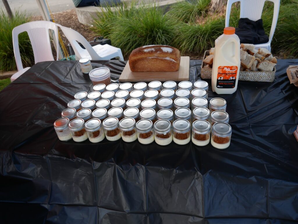 Rows of small jars with silver lids on a black-covered table beside a loaf of bread and a bottle of cream outdoors.