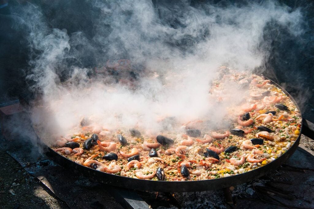 A large paella cooking outdoors over flame, with rice, peppers and chicken