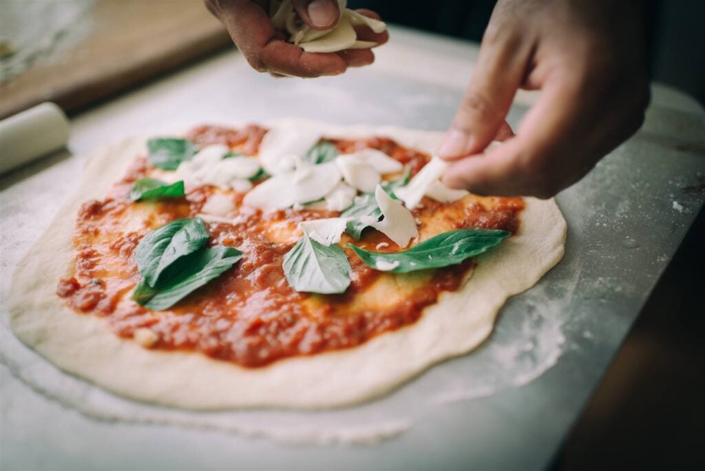 Hands topping a hand-stretched pizza with fresh basil and mozzarella at a Fleurieu Food and Wine pizza class