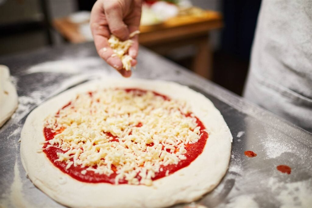 Cheese being scattered over tomato sauce on a hand-stretched pizza base at a cooking class