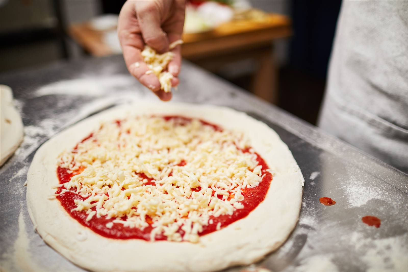Cheese being scattered over tomato sauce on a hand-stretched pizza base at a cooking class