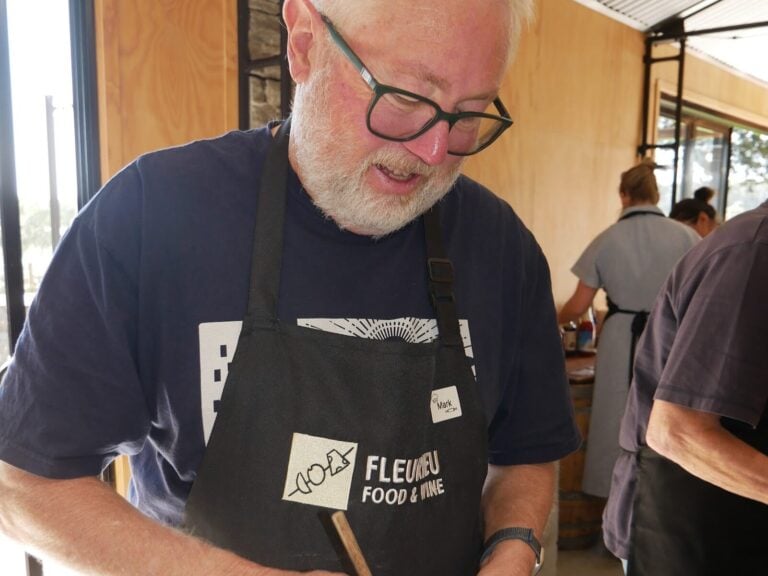 A man in a black apron stirs flour and egg in a metal bowl with a wooden stick while others work behind him.
